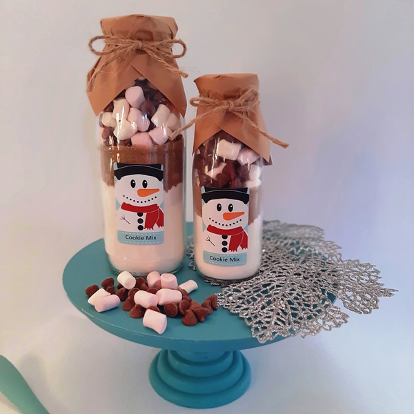 Two bottles of cookie mix with snowman labels on a blue stand with marshmallows and chocolate pieces.